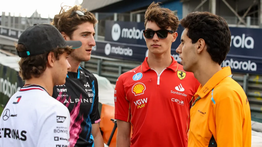 Quatro jovens pilotos estão reunidos em uma corrida de Fórmula 1. A composição da imagem mostra os quatro jovens, de frente para a câmera, em uma disposição informal. Eles parecem estar em uma conversa, possivelmente em um pit stop ou área de apoio. Os indivíduos estão vestidos com uniformes de equipe esportiva, com cores e logos que sugerem sua afiliação a diferentes equipes de Fórmula 1, como Alpine, Ferrari e McLaren, entre outras.