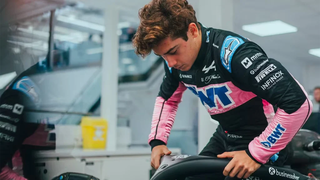 Uma imagem mostra um jovem inclinado sobre um assento de carro. A composição da imagem centra-se no jovem, com o seu corpo inclinado para a frente. Ele está em primeiro plano, enquanto o fundo apresenta elementos de um ambiente de preparação de veículos, como máquinas e paredes claras. O enquadramento é fechado, concentrando-se na figura principal e na sua ação.
