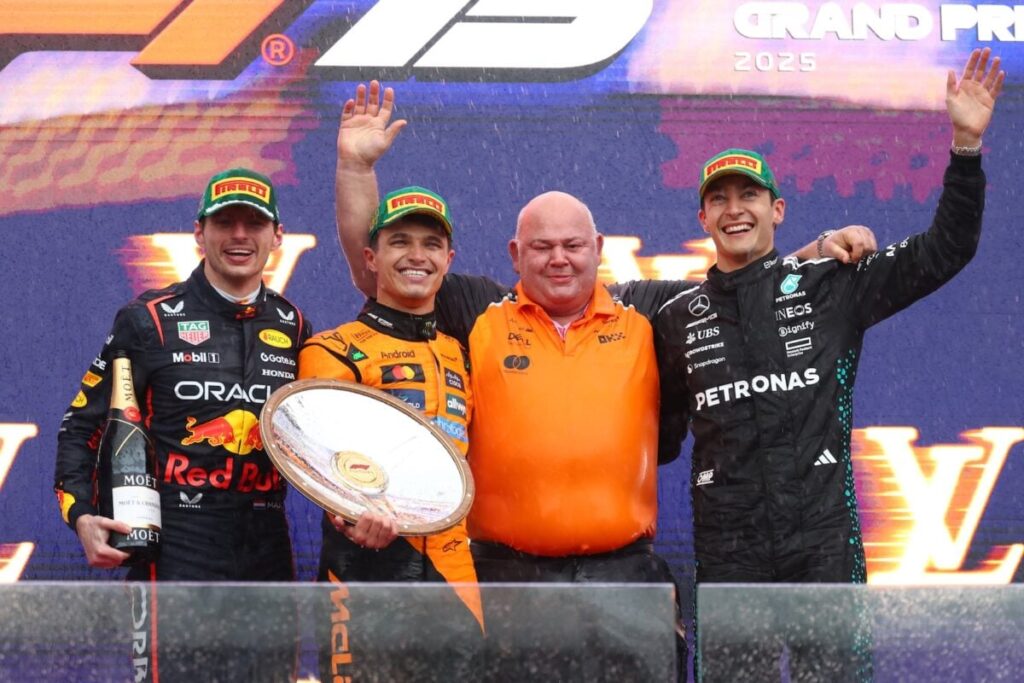 Três pilotos de Fórmula 1 e um membro da equipe McLaren posam no pódio, após uma corrida, com expressões de alegria e realização. O pódio é composto por três degraus, com os pilotos em posições diferentes e com acessórios como champanhe e troféus. Há uma hierarquia visual, com os pilotos da frente sendo mais destacados. Os pilotos exibem sorrisos. O enquadramento é completo, mostrando todos no pódio.