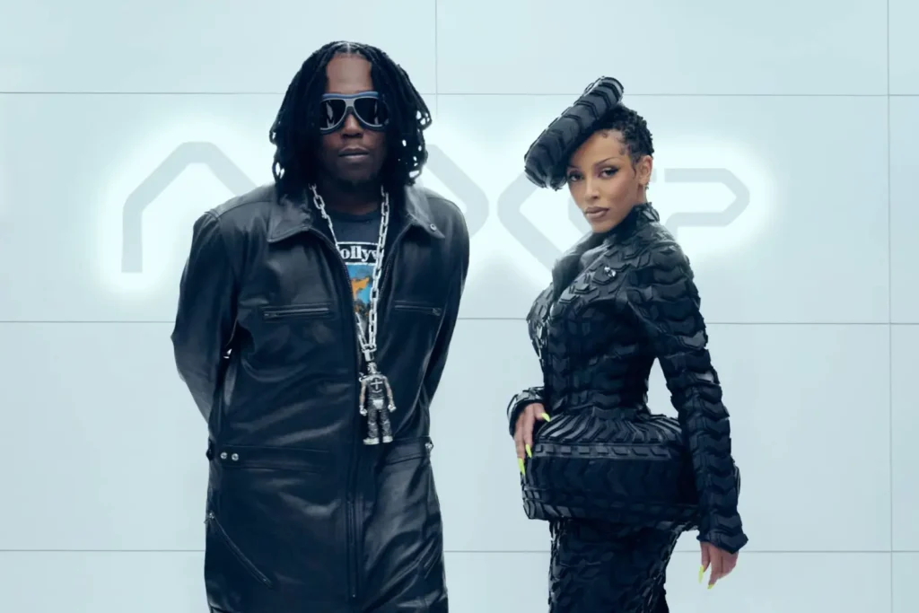Uma cena de videoclipe contemporâneo apresenta dois indivíduos, um homem e uma mulher, Don Toliver e Doja Cat, posicionados contra um fundo de painéis cinza. A cena é composta com as duas figuras posicionadas centralmente, de frente para a câmera. O homem, à esquerda, está ligeiramente à frente, vestindo um macacão de couro preto e óculos de sol estilo aviador. A mulher, à direita, veste uma jaqueta preta esculpida, com um design único.
