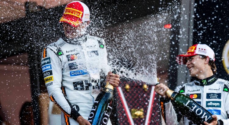 A composição da imagem mostra dois homens, pilotos de corrida, em meio a uma chuva de champanhe. Eles estão em primeiro plano, com uma grande quantidade de champanhe sendo derramada em suas roupas e ao redor deles. Os dois homens estão posicionados um ao lado do outro, e cada um segura uma garrafa de champanhe e está comemorando a vitória. O fundo mostra uma plateia e vários detalhes em um cenário de corrida, com uma atmosfera de entusiasmo e celebração.