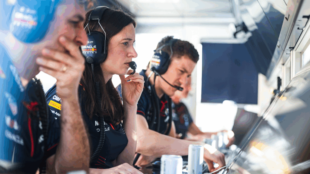 Vários membros de uma equipe de corrida de Fórmula 1, possivelmente da equipe Red Bull Racing, estão concentrados em uma sala de controle, com fones de ouvido e aparentemente focados em suas tarefas. A composição da cena mostra uma série de indivíduos, aparentemente envolvidos em uma atividade colaborativa em uma sala de controle. Os indivíduos estão posicionados próximos uns aos outros, possivelmente em uma área de trabalho de equipe, interagindo por meio de comunicações, sugerindo o gerenciamento de informações críticas durante uma corrida de Fórmula 1. O enquadramento é fechado, focando na interação entre as pessoas, e a profundidade de campo é rasa, com um foco principal nas expressões e ações das pessoas no primeiro plano. Há uma sensação de intensidade e concentração no ambiente, devido à postura, expressões e a atenção focada.