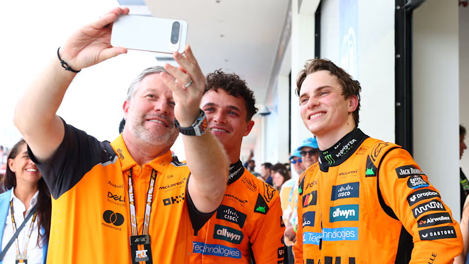 Fotografia dos pilotos Lando Norris e Oscar Piastri em seus macacões cheio de patrocinadores nas mesmas cores da equipe britânica, laranja papaia e preto, juntos de Zak Brown, diretor executivo da equipe, vestido em uma blusa das mesmas cores para uma foto após a corrida.