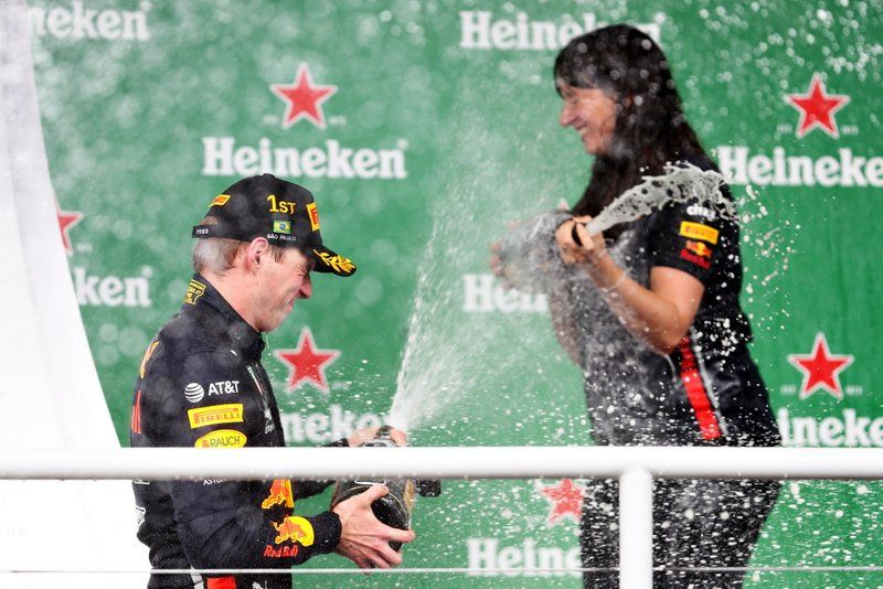 A composição da imagem mostra dois indivíduos em primeiro plano, provavelmente participantes de uma corrida de automóveis, em meio a uma neblina de champanhe. Eles estão em posições de celebração, aparentemente envolvidos em um momento de comemoração, em que o champanhe é borrifado sobre eles. O fundo exibe um padrão de cores verdes e logos de empresas, como Heineken e AT&T, sugerindo um local de evento público. Os participantes estão usando roupas escuras, possivelmente uniformes de corrida ou roupas específicas do evento. A imagem foca na interação dos indivíduos, enfatizando a celebração e os detalhes da névoa de champanhe no primeiro plano.