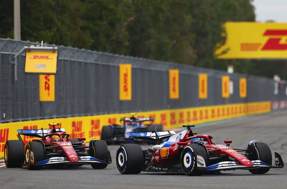 Em primeiro plano, ambos carros da Ferrari em sua tradicional cor vermelha sangue mas com espaços na asa dianteira, pneu, asa traseira e parte de cima da abertura do motor em cores azul e branca, ultrapassando o carro azul escuro e preto da equipe Williams, localizada em segundo plano.