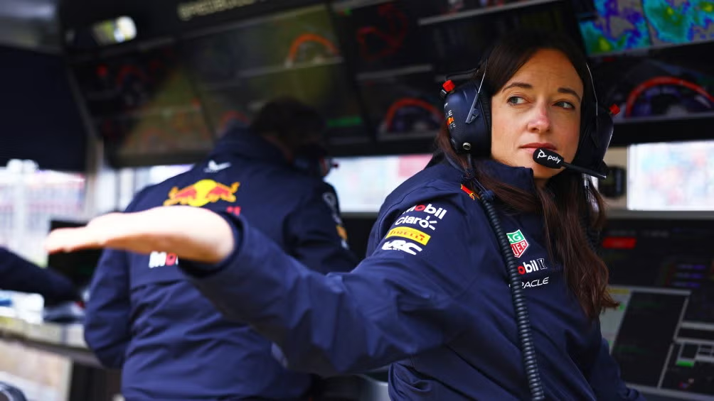 A imagem mostra uma mulher de meia-idade com cabelo castanho escuro, usando fones de ouvido e um uniforme de uma equipe de corrida, possivelmente um uniforme de trabalho. Ela está posicionada de forma que seu corpo está ligeiramente voltado para a esquerda, enquanto seu braço direito está estendido. A postura sugere que ela está em uma postura ativa e comunicativa. O foco principal está na figura da mulher, e o fundo mostra monitores e equipamentos de controle de uma equipe de corrida. O fundo é composto de várias telas de computador e equipamentos de monitoramento que indicam um ambiente técnico e de controle.